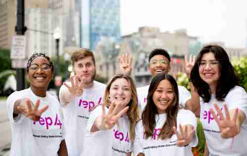 Students with Rep4 t-shirts smile and make a number 4 sign with their fingers in a downtown setting